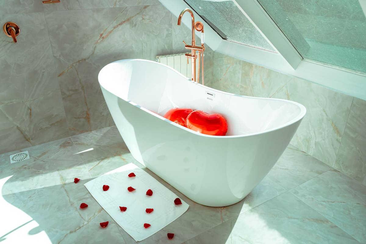 A modern bathroom with a freestanding bathtub, a window above the sink, and rose petals on the floor.
