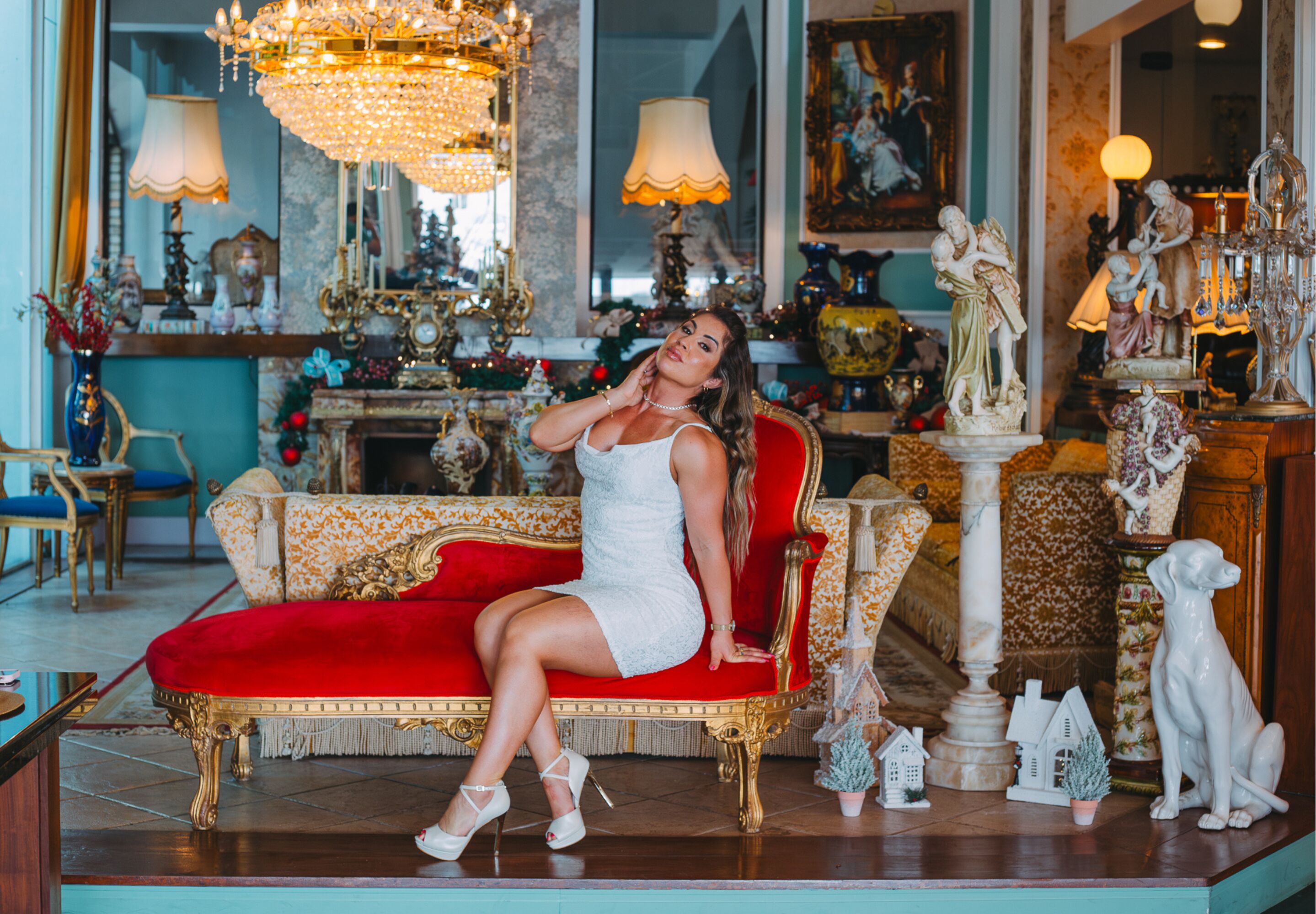 A woman seated on a red and gold ornate sofa in a richly decorated room with chandeliers, statues, and various decorative items.