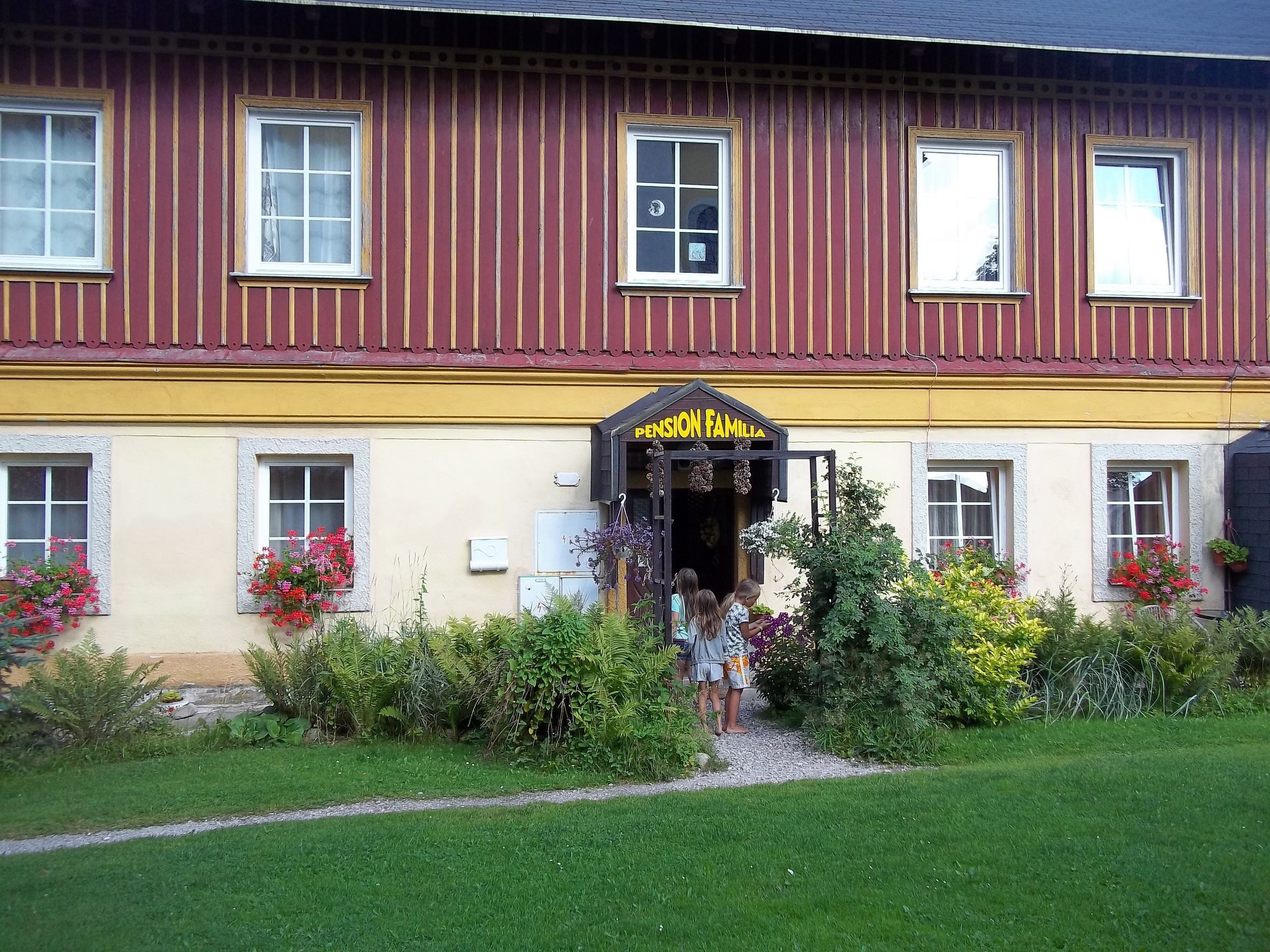 A building with a yellow and red facade, a sign reading "PENSION FAMILIA," and a garden with various plants and flowers.