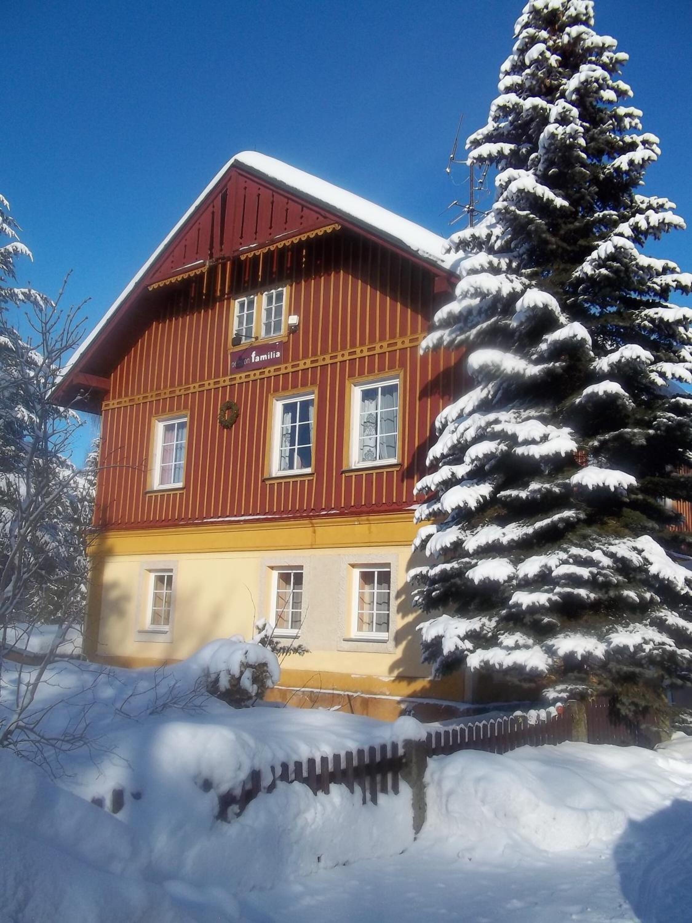 A two-story building with a red and yellow facade, a sign reading "Hotel Familia," and a snow-covered tree in front.