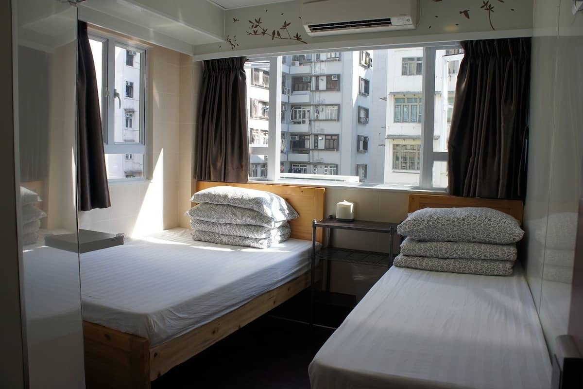 Une chambre d’hôtel avec deux lits, une fenêtre avec des rideaux, une table de chevet avec une bougie et une vue sur les bâtiments environnants.
