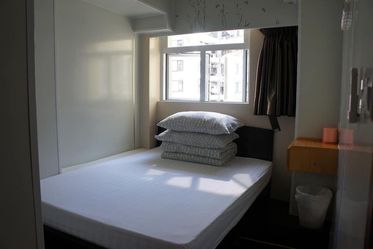 Une petite chambre avec un lit simple, une fenêtre, une table de chevet et une poubelle.