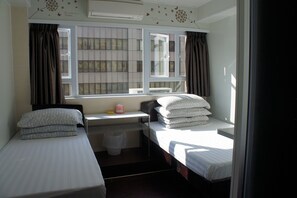 Una habitación pequeña y funcional con dos camas individuales, una ventana con cortinas, una mesita con una caja de pañuelos y un aire acondicionado instalado en la pared.