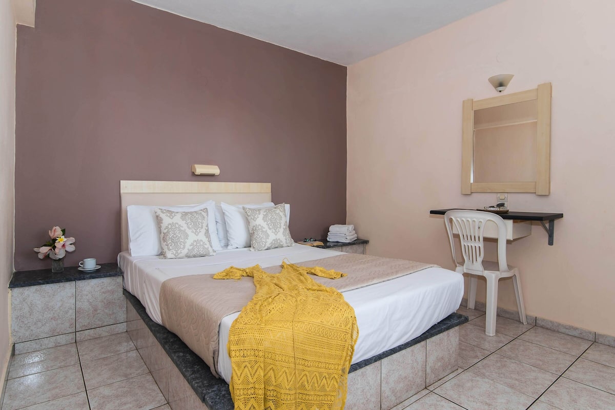 A hotel room with a bed, a desk, a chair, a mirror, and a vase with flowers.