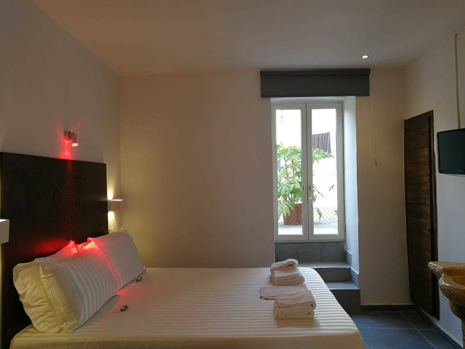 A hotel room with a bed, a TV, a window with a view of the outside, and a red light on the wall.