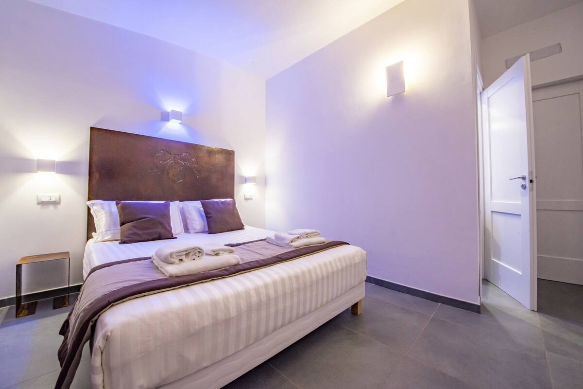 A modern hotel room with a neatly made bed, a bedside table, and a wall-mounted light fixture above the bed.