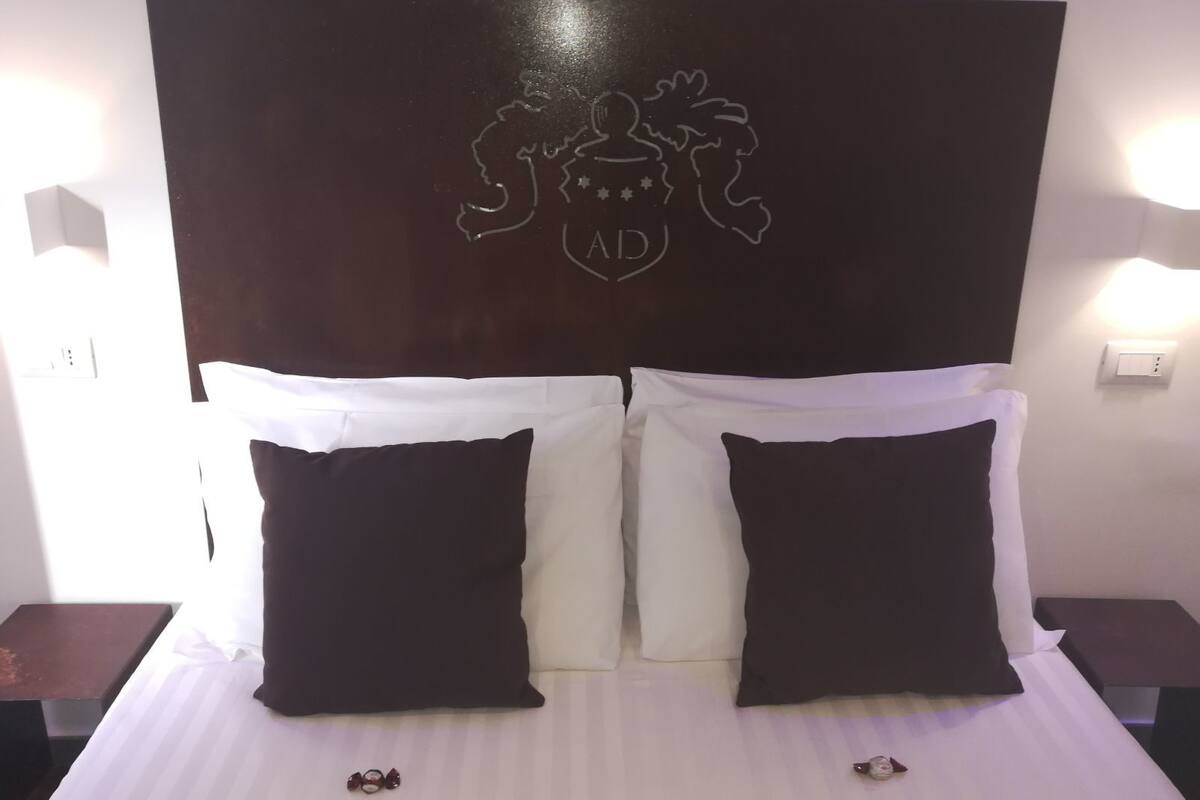 A hotel room with a large bed, white bedding, and a dark wooden headboard featuring a crest.
