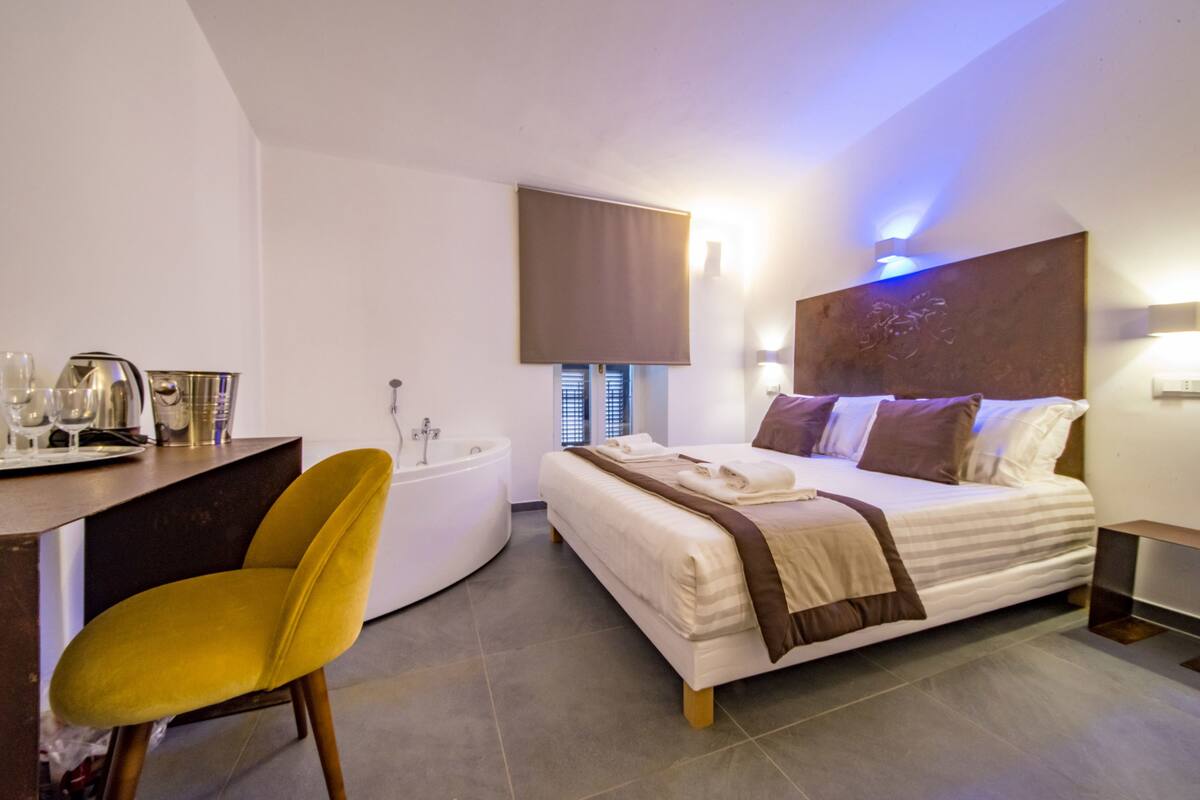 A modern bedroom with a bed, a desk, a chair, a bathtub, and a window with blinds.
