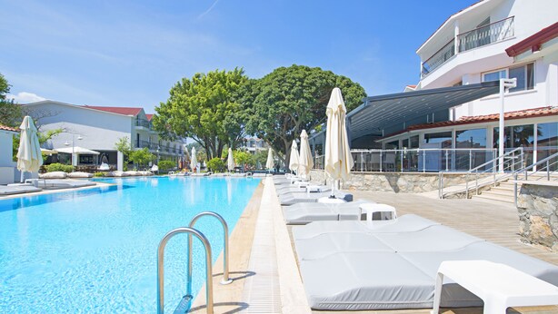 Zona de piscina con sillones de descanso y sombrillas, cielo despejado y un edificio con balcón.