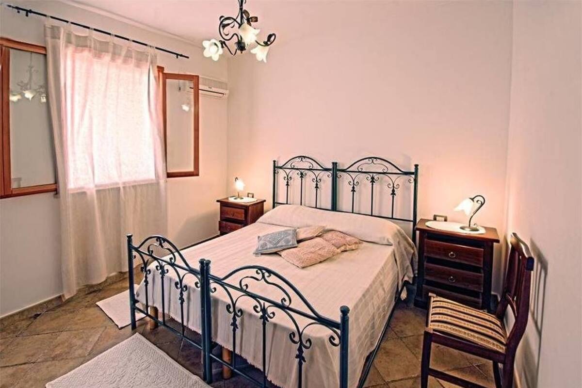 Une chambre à coucher avec un lit en métal, une chaise en bois, une table de chevet avec une lampe et une fenêtre ornée de rideaux légers.