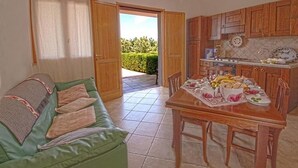 A cozy living room with a green sofa, a dining table set with food, and a kitchen with wooden cabinets. - Agriturismo Sa Suerera (Olmedo)