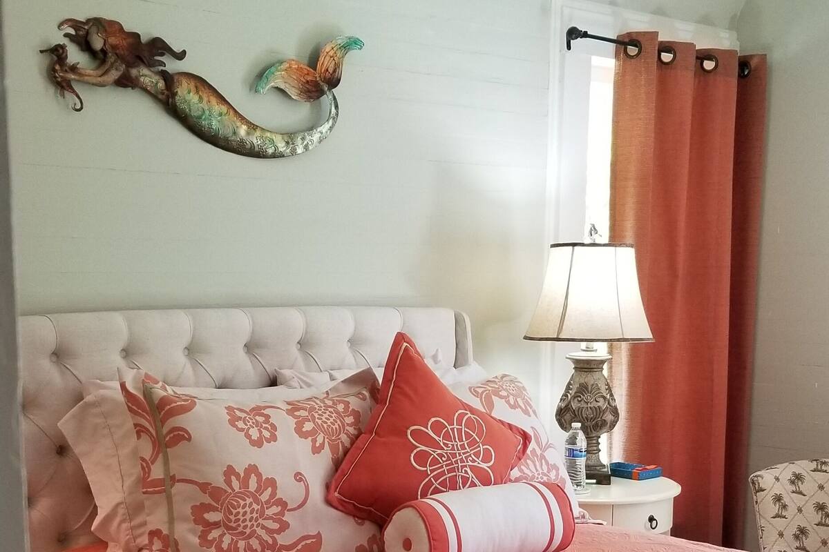 A bedroom with a tufted headboard, floral-patterned pillows, a red blanket, a decorative wall piece, a bedside lamp, and a patterned rug.