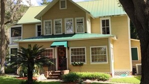 Une maison jaune à deux étages, avec un toit vert, un porche à l’avant et un petit jardin agrémenté d’un palmier.
