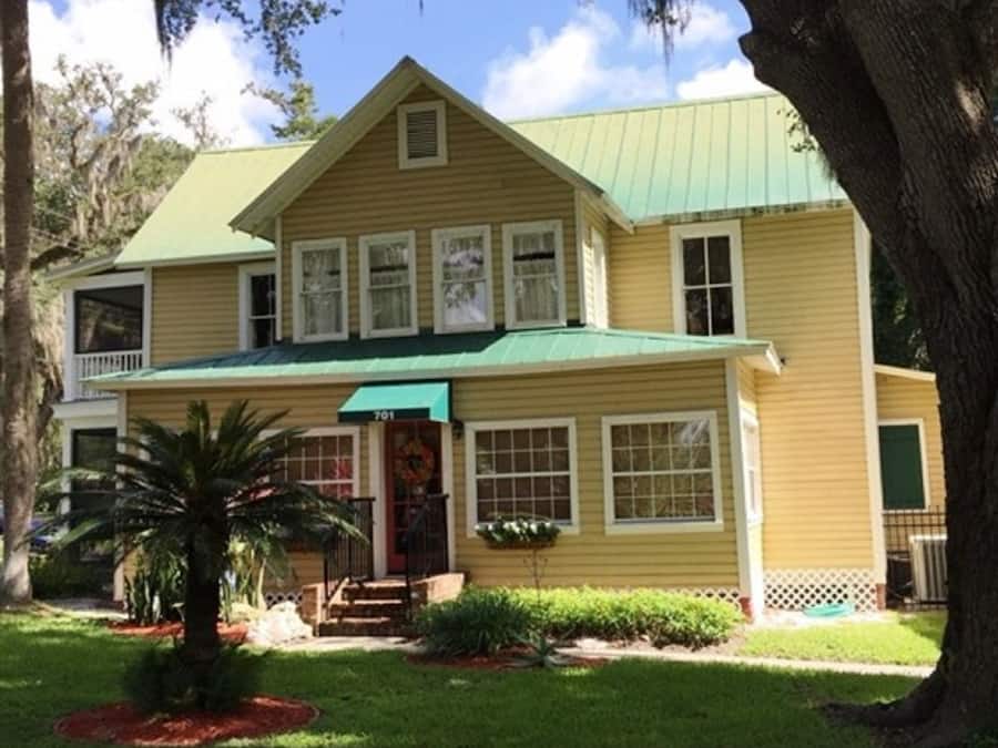 Una casa amarilla de dos pisos con techo verde, un porche delantero y un pequeño jardín con una palmera.