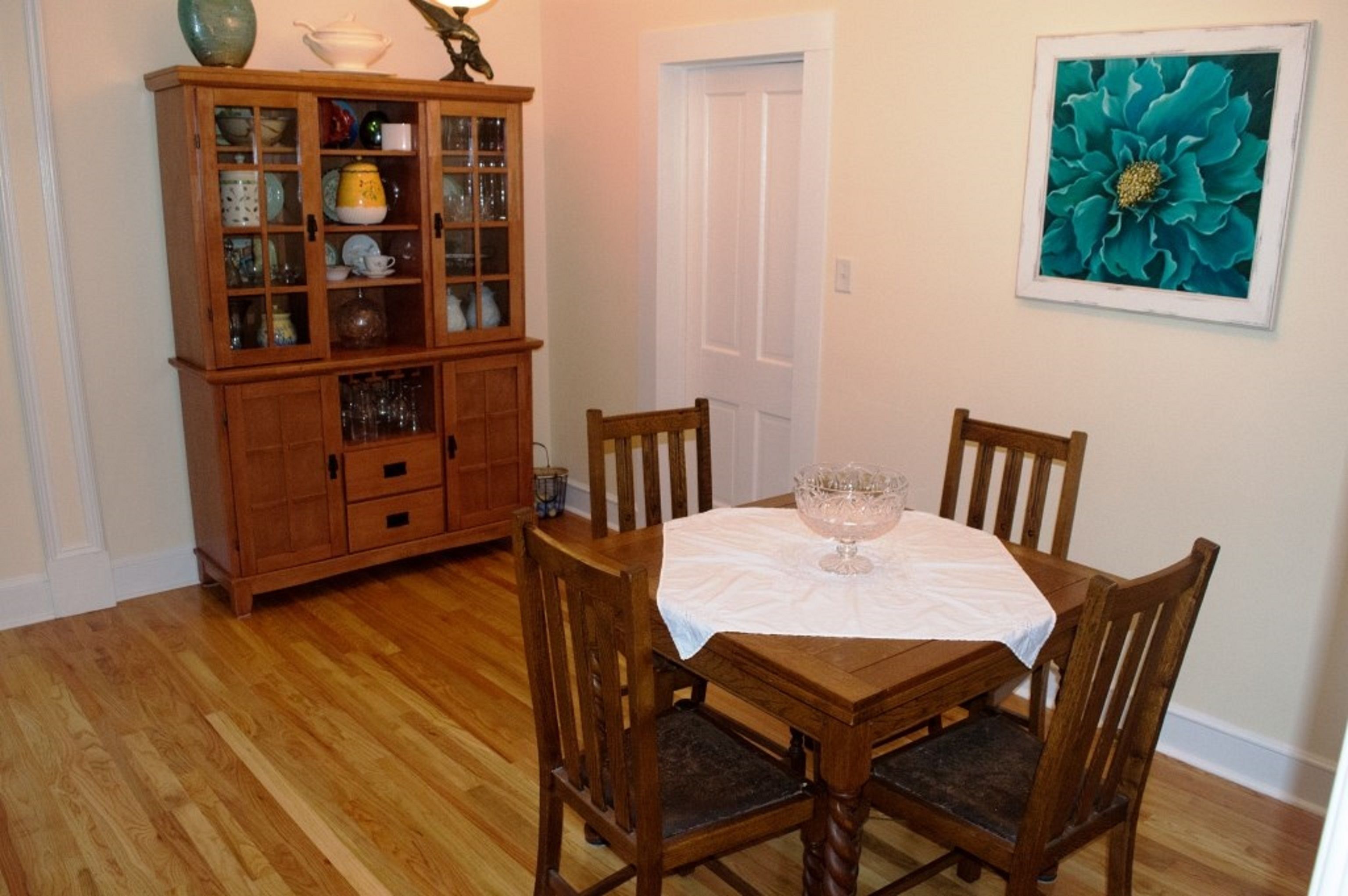 Un coin repas avec une table et des chaises en bois, une vitrine contenant de la vaisselle et un tableau floral encadré accroché au mur.