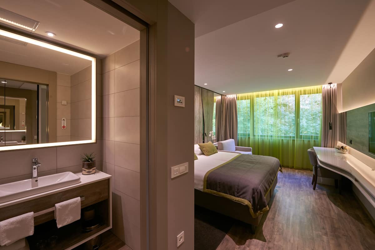Una habitación de hotel moderna con una cama grande, una mesita de noche, un escritorio y un baño con lavabo y espejo.