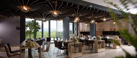 Un restaurante moderno con una decoración contemporánea, que presenta un área de comedor central con mesas y sillas, rodeado de grandes ventanales con vistas al exterior.