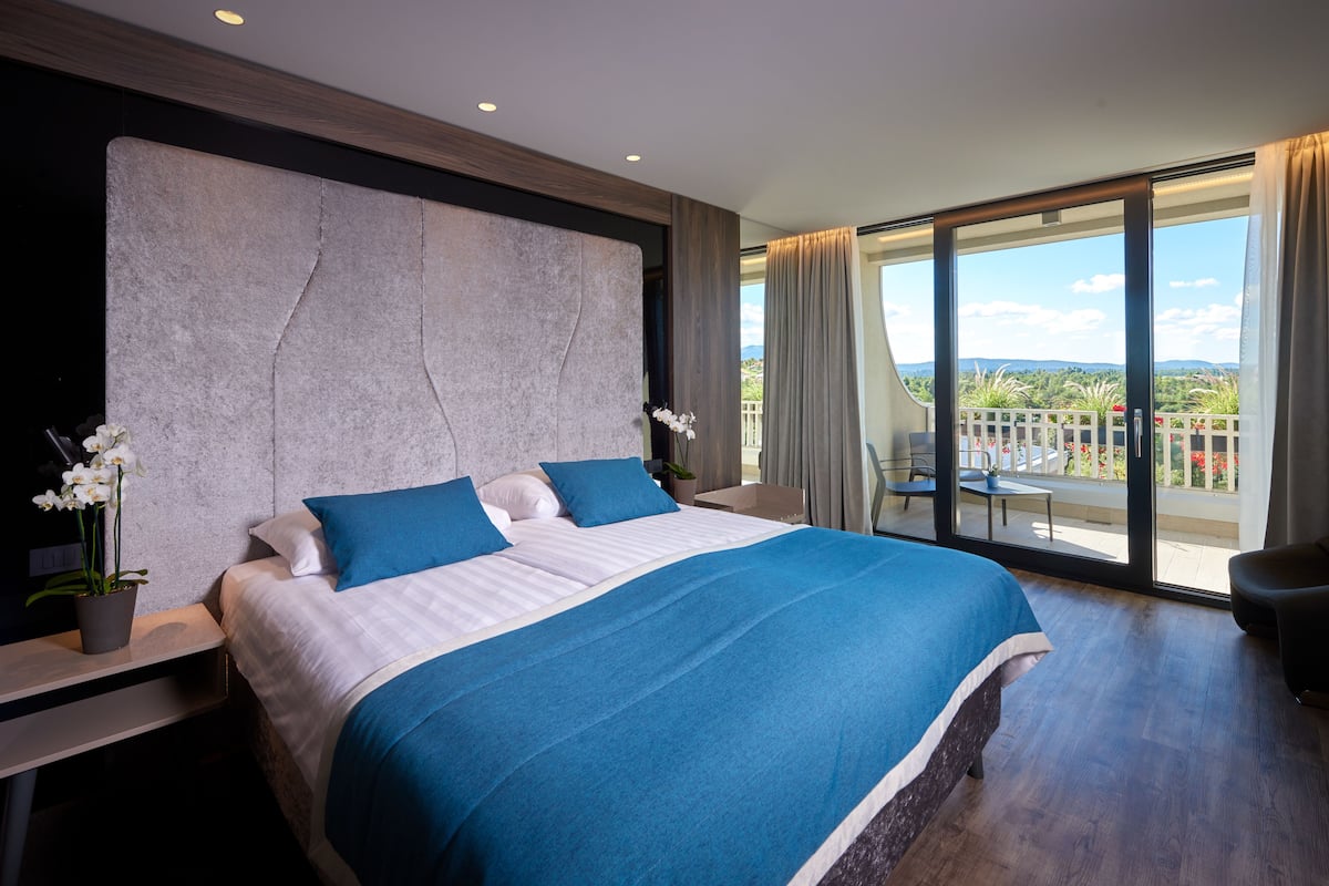 Habitación de hotel moderna con una cama grande, una mesita de noche, una planta en maceta y un balcón con vistas.
