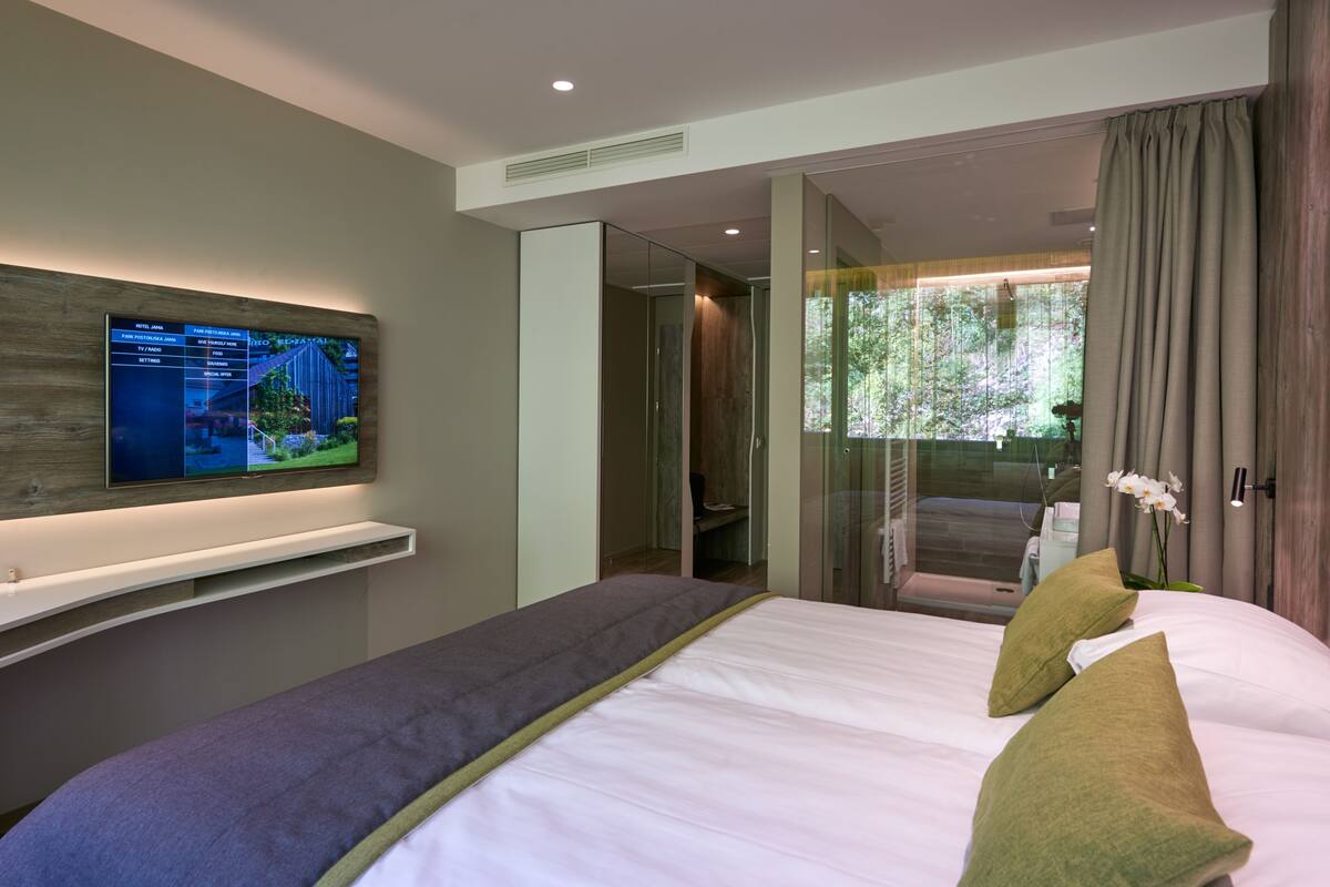 Una habitación de hotel moderna con cama, televisor montado en la pared, puerta de cristal que da al baño y vistas a zonas verdes.