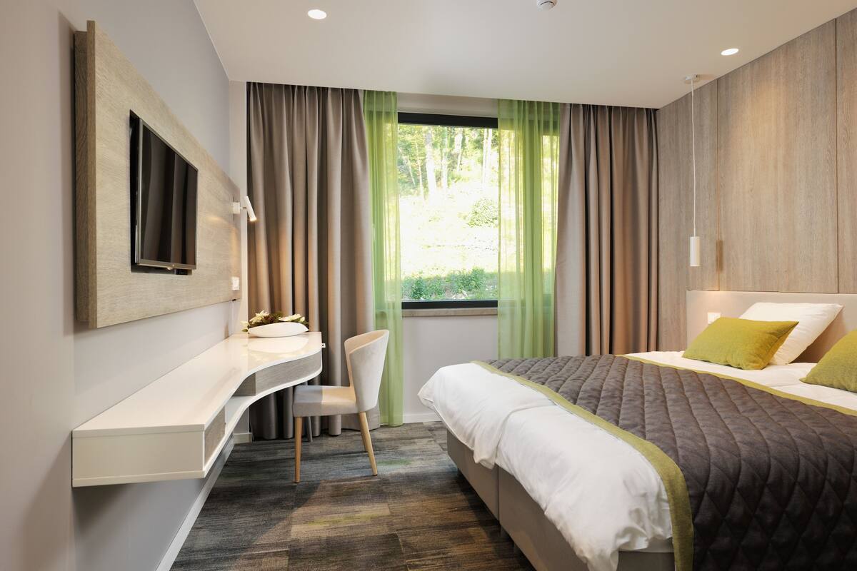 Una habitación de hotel moderna con una cama, un escritorio con silla, un televisor en la pared y una ventana con cortinas.