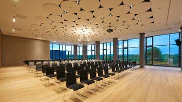 Ein Konferenzraum mit Reihen von Stühlen, großen Fenstern und einem modernen Deckenstil.