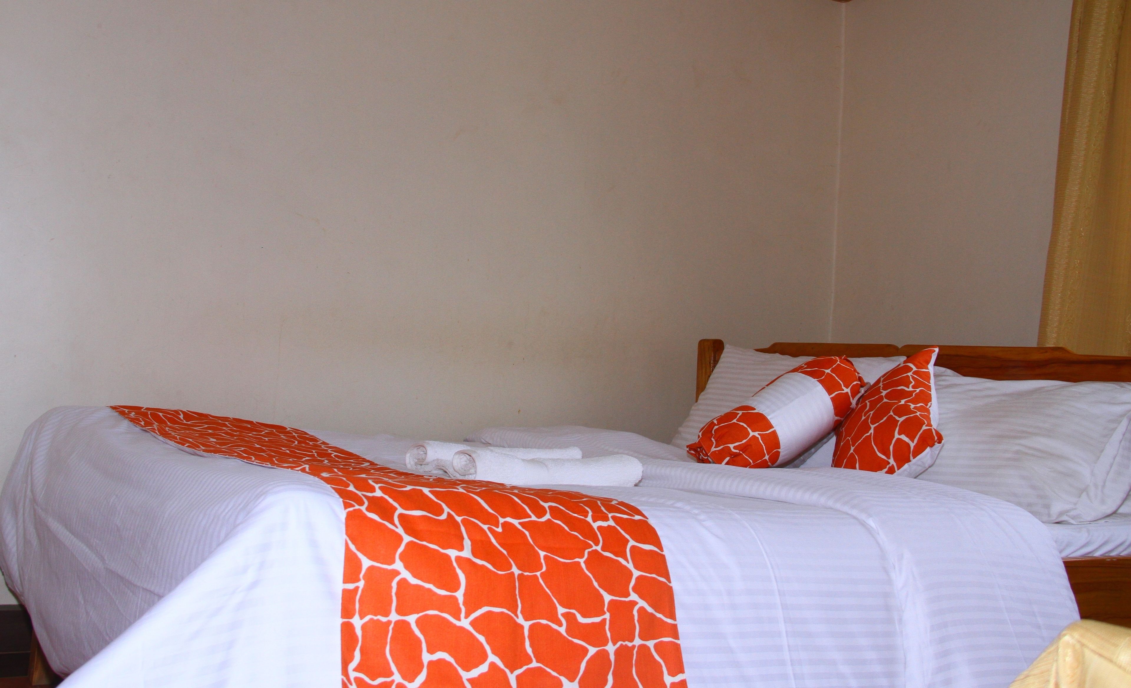 A neatly made bed with white and orange bedding, a wooden headboard, and a plain wall with a curtain.