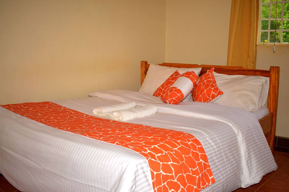 Una cama bien hecha con una colcha blanca y naranja estampada, dos almohadas con estampado de jirafa y un par de toallas blancas dobladas.