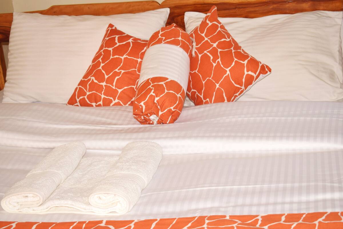 Una cama bien hecha con almohadillas blancas y naranja estampadas, una colcha a rayas blancas y dos toallas blancas enrolladas.