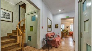 Un pasillo con suelo de madera, una escalera, una puerta verde, un sofá rosa y una pequeña mesa con un jarrón.