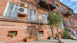 Un edificio de ladrillo rojo con varias ventanas, algunas protegidas por rejas metálicas, y un balcón. Hay plantas en macetas a nivel de la calle y una mesa de metal.
