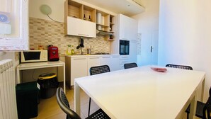 A modern kitchen with white cabinets, a microwave, and a stove. There is a dining table with chairs and a clock on the wall. - Liola Home (Palermo)