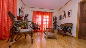 A room with wooden flooring, a wooden chair, a wicker sofa, a small table, and a window with red curtains.