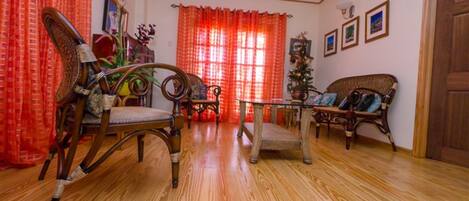 A room with wooden flooring, a wooden chair, a wicker sofa, a small table, and a window with red curtains.
