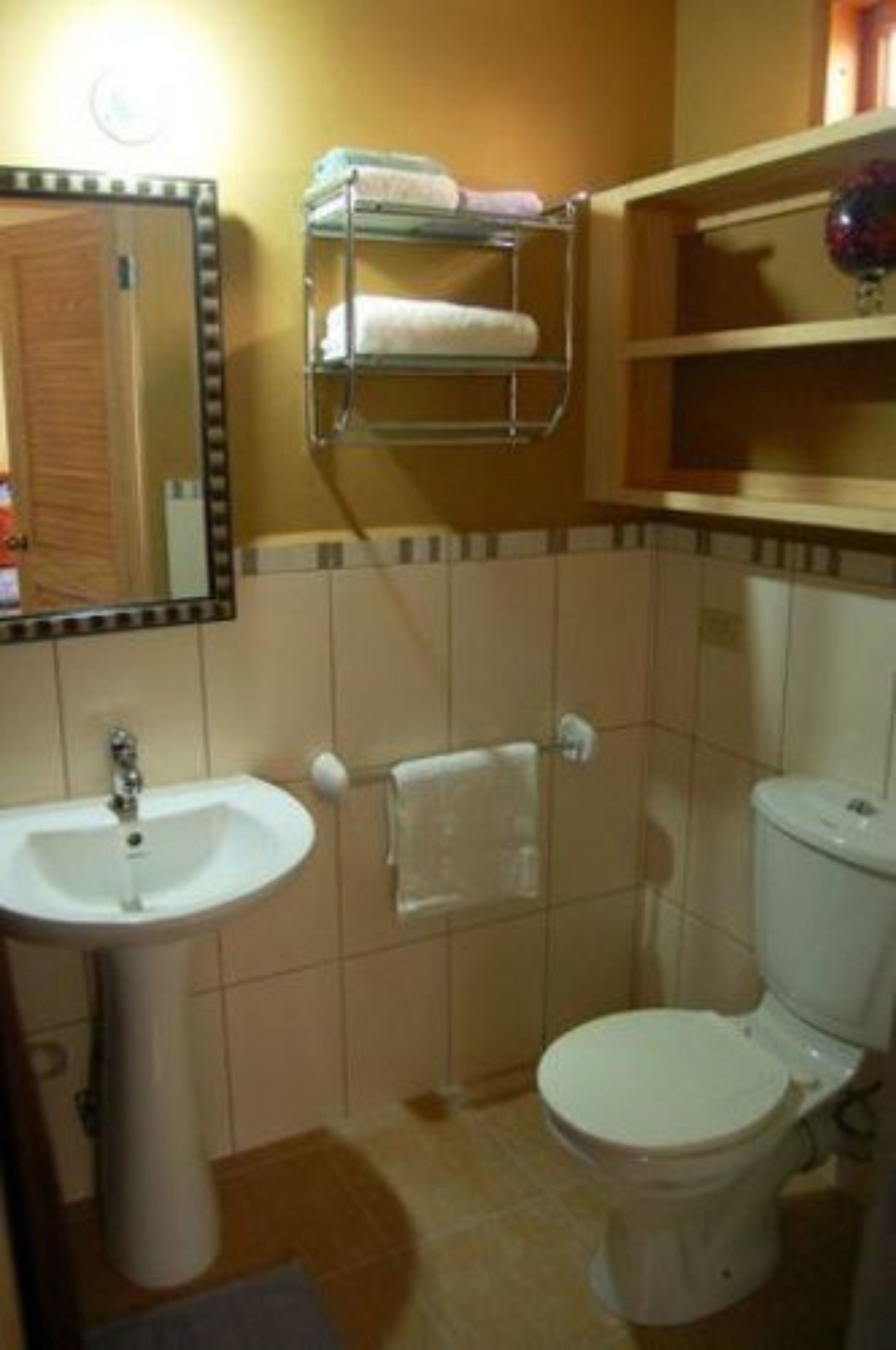 A bathroom with a sink, toilet, and a mirror with lights. There is a towel rack above the toilet and a shelf with toiletries.