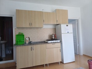 A kitchen with wooden cabinets, a gas stove, a white refrigerator, and a green plastic basket. - Elite Apart Pansiyon (Gökçeada)
