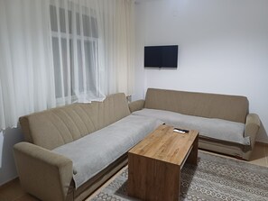 Una sala de estar con un sofá, una mesa de centro, un televisor montado en la pared y cortinas traslúcidas.