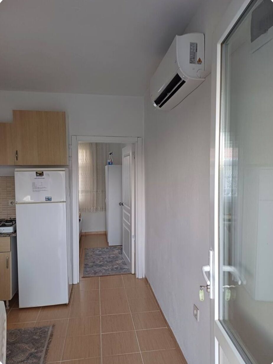 A compact kitchen with a refrigerator, an air conditioner, and a door leading to a hallway.
