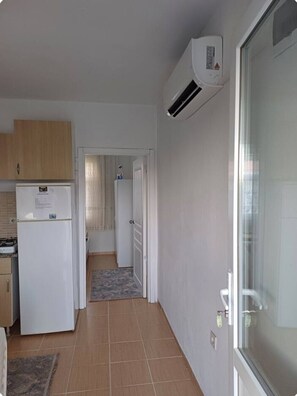 A compact kitchen with a refrigerator, an air conditioner, and a door leading to a hallway. - Elite Apart Pansiyon (Gökçeada)