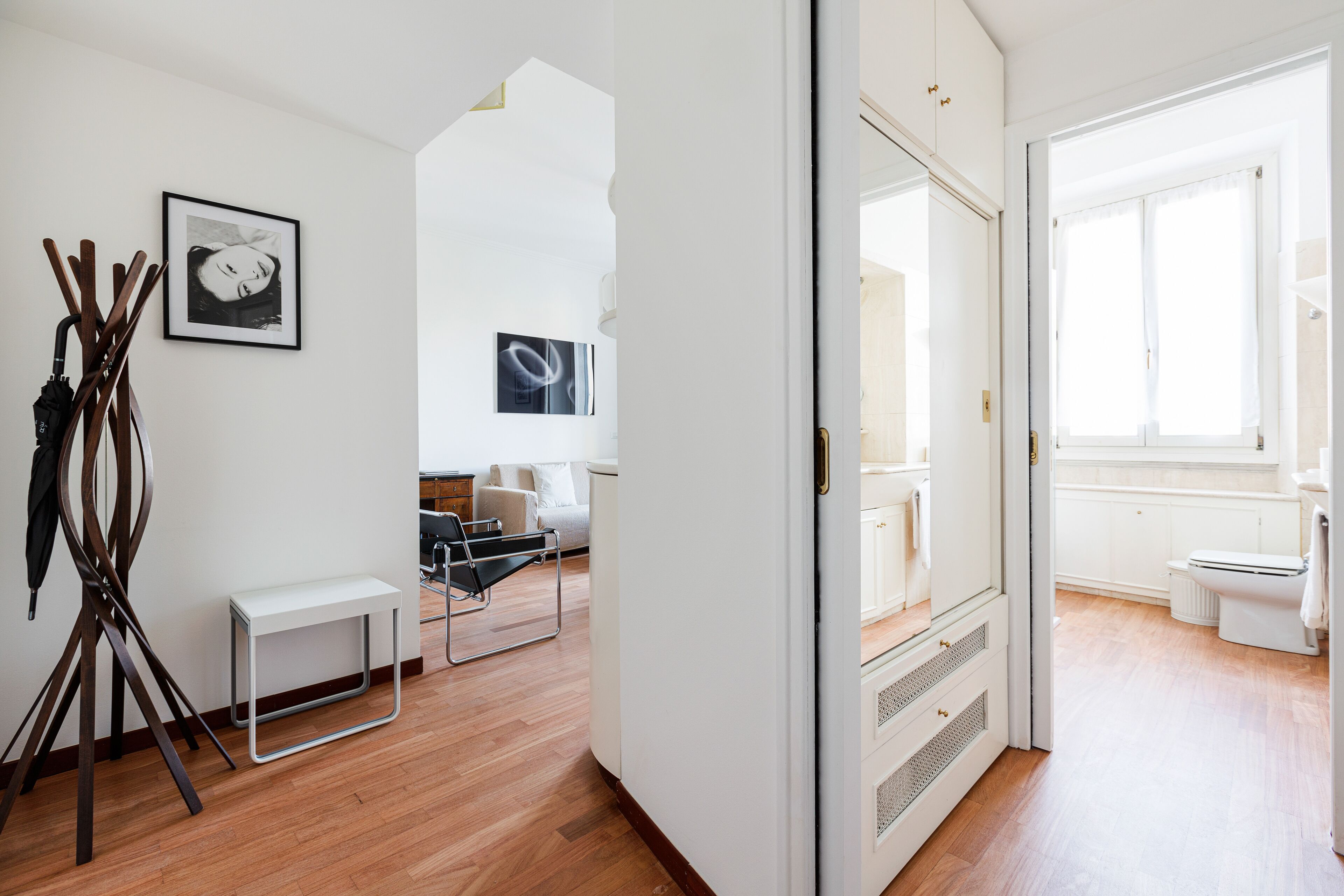 A modern, well-lit room with wooden flooring, a white bench, a black chair, and a coat rack.