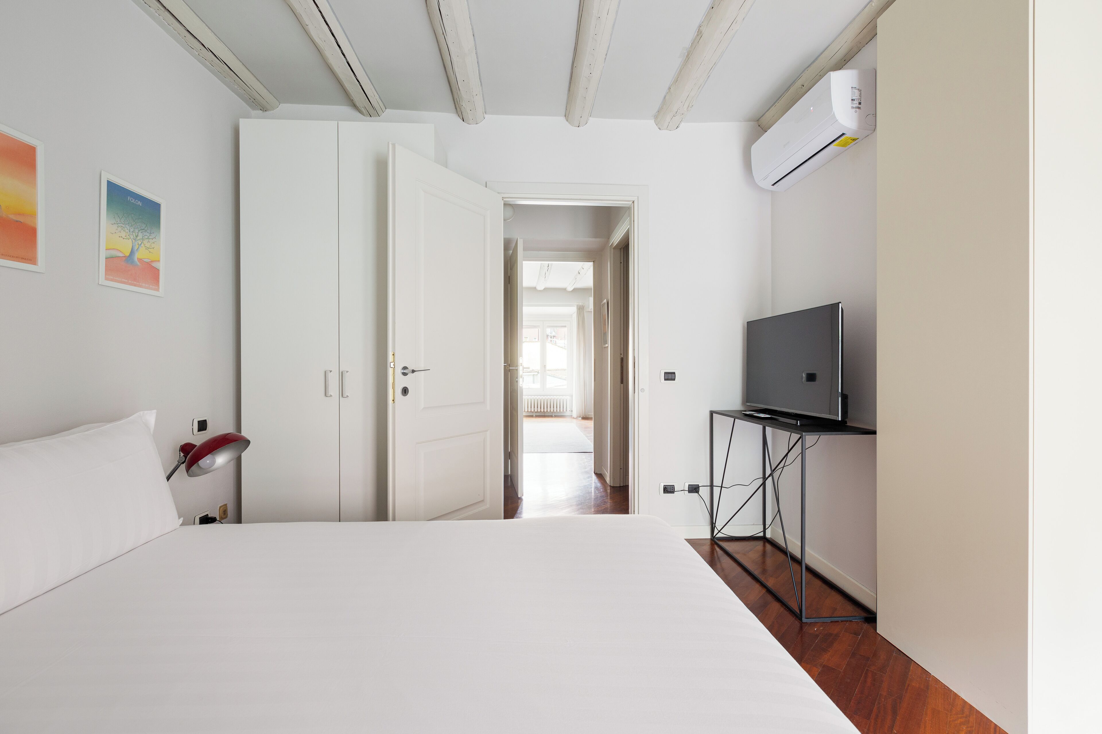 A bedroom with a bed, a desk, a TV, and a door leading to another room.