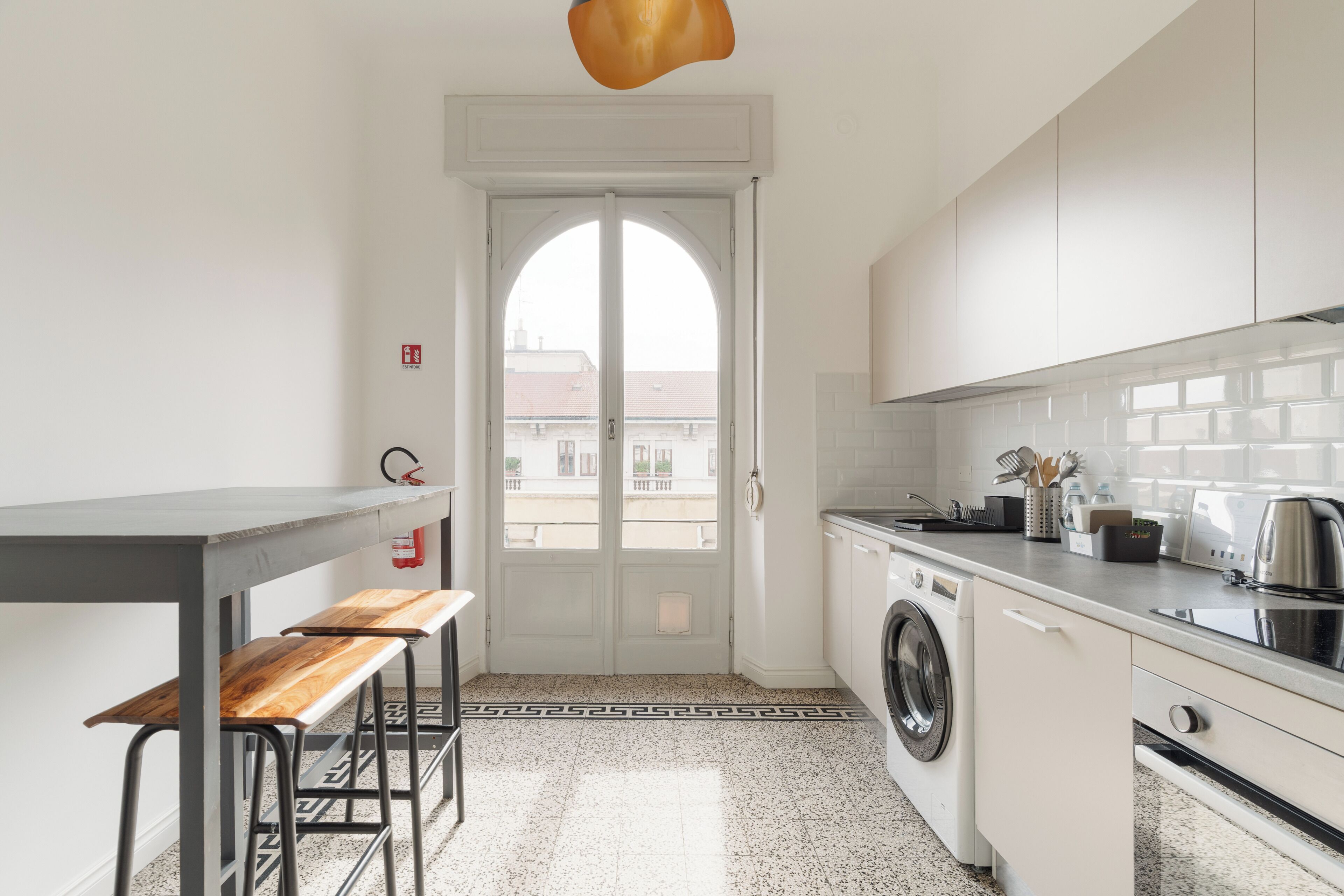Une petite cuisine équipée d’un lave-linge, d’un évier et d’une fenêtre donnant sur un immeuble.