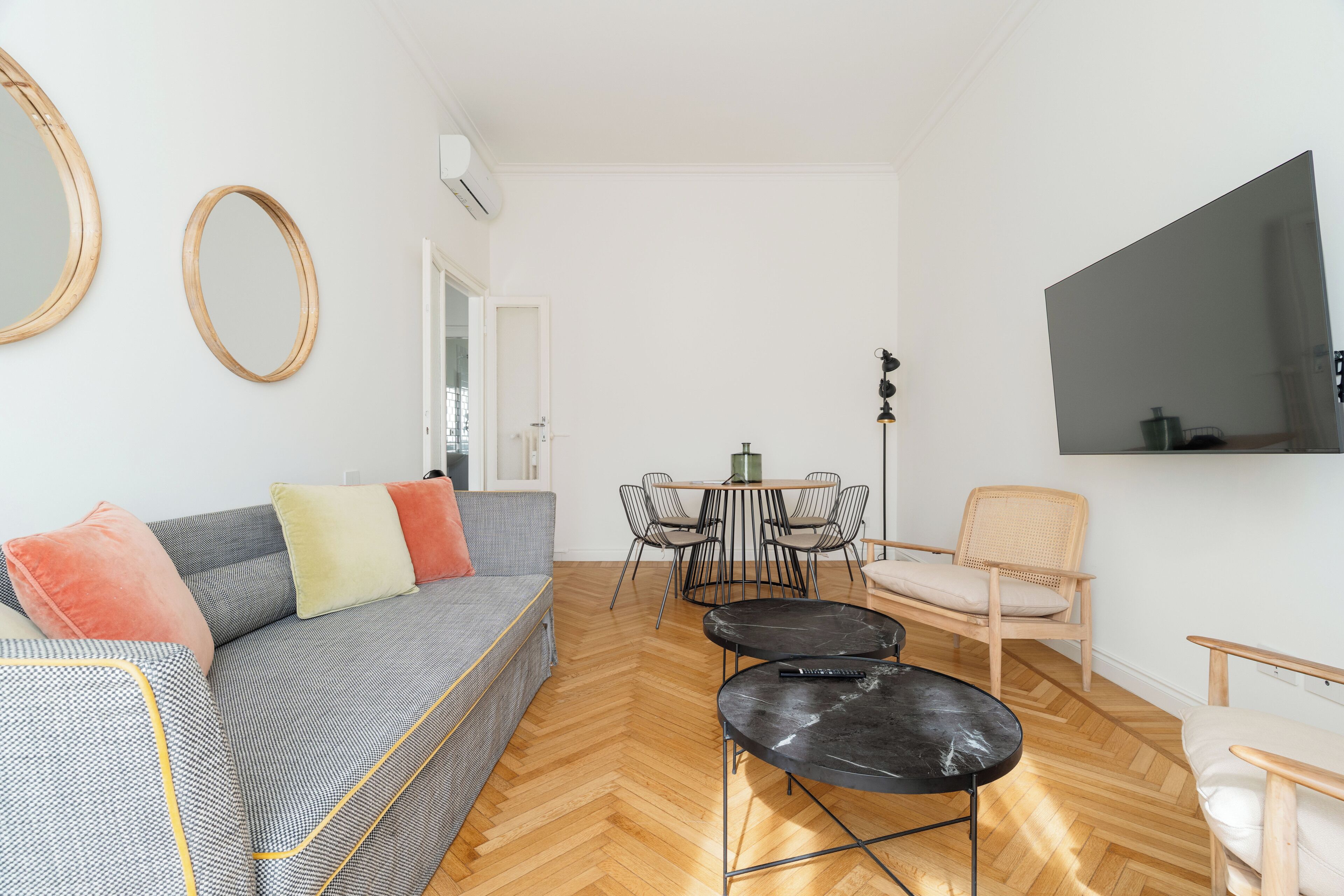 Un salon avec un canapé, une table à manger et des chaises, un téléviseur fixé au mur et un plancher en bois.