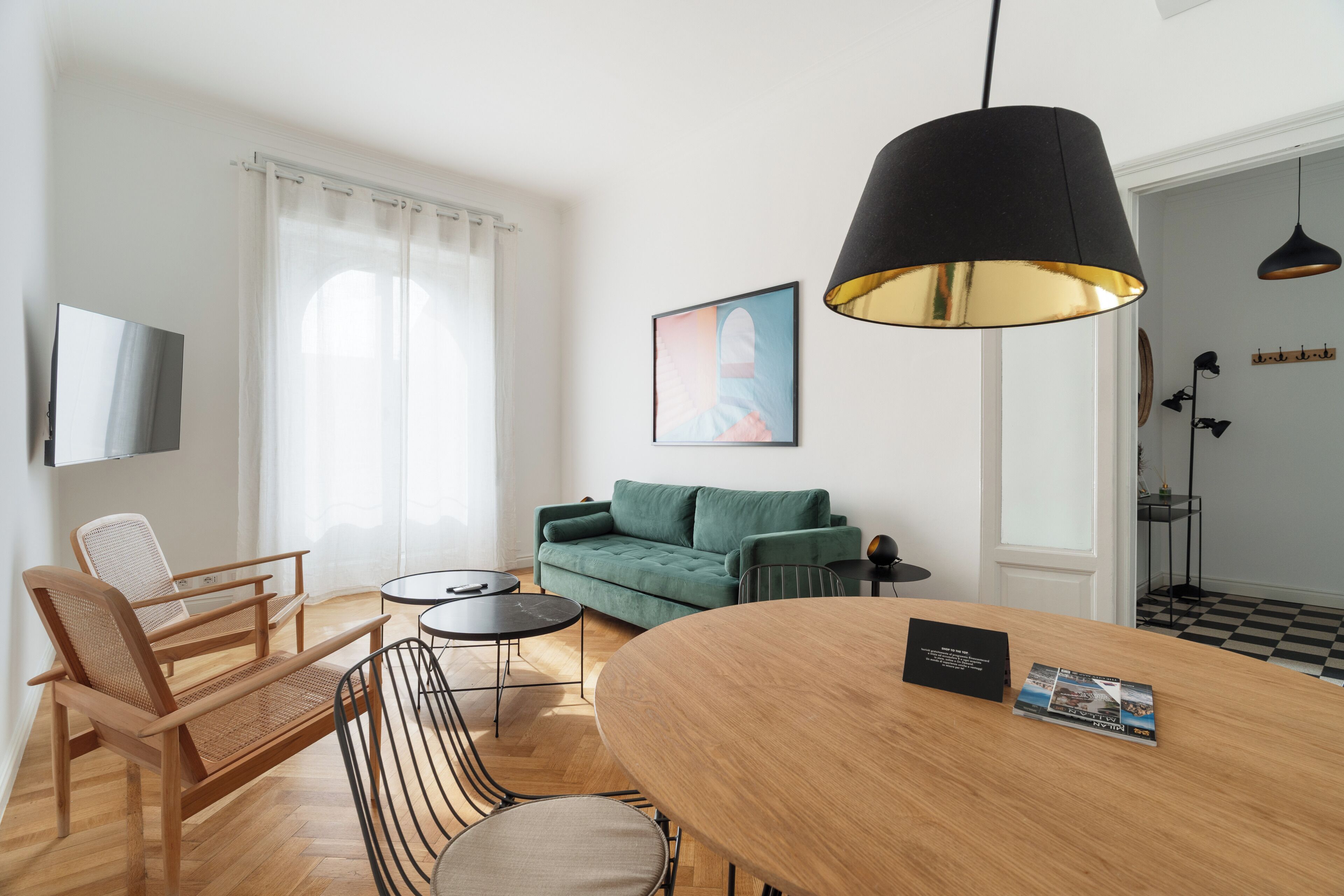 A modern living room with a wooden dining table, a green sofa, a TV mounted on the wall, and a large black pendant light.
