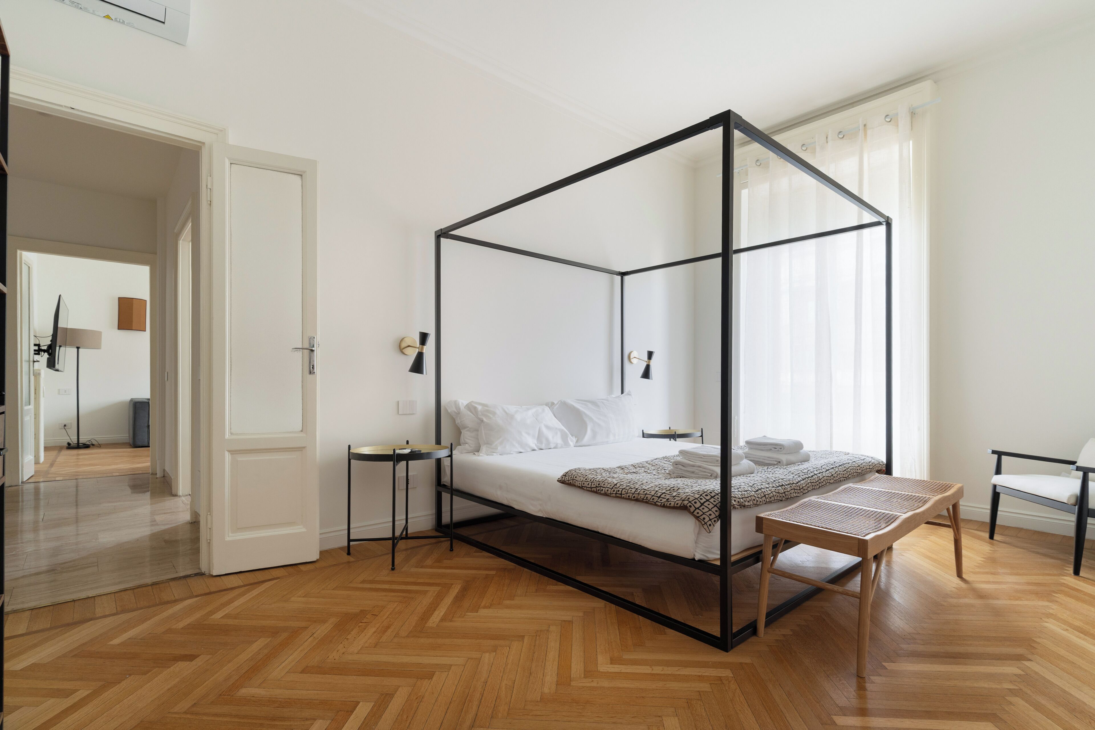 Una cama con dosel de madera, una mesita de noche, una silla y vista a una habitación con una lámpara y una ventana.