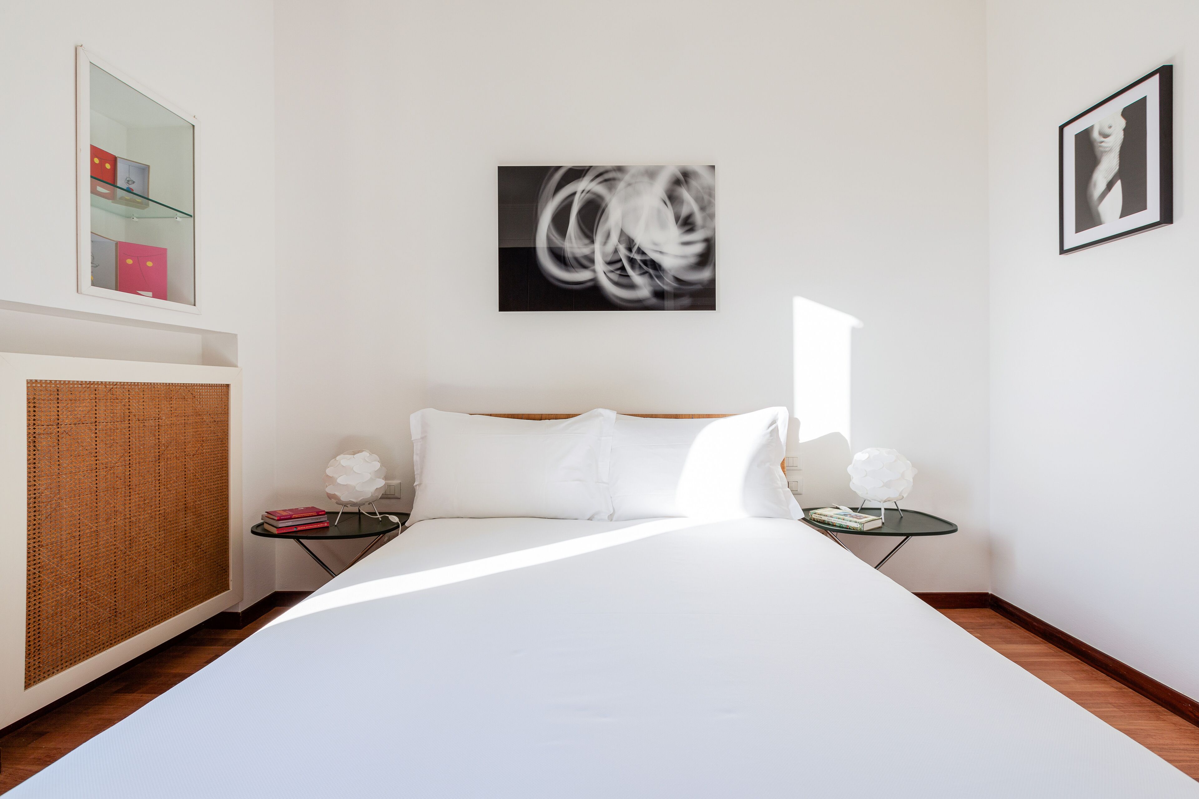 A modern bedroom with a large bed, a small bedside table, a mirror, and a wall-mounted abstract painting.