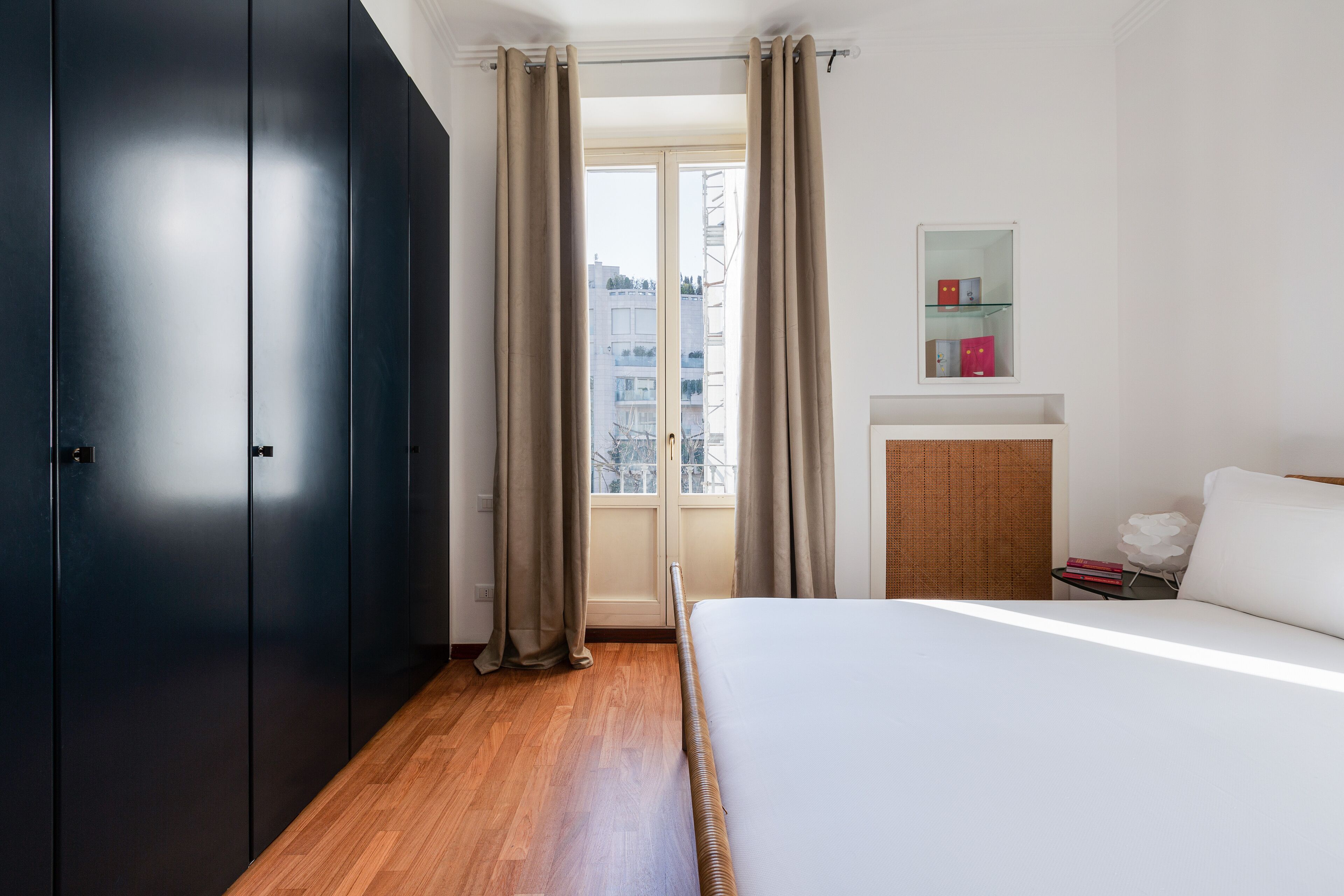 A bedroom with a bed, a wardrobe, a window with curtains, a bedside table with books, and a mirror on the wall.