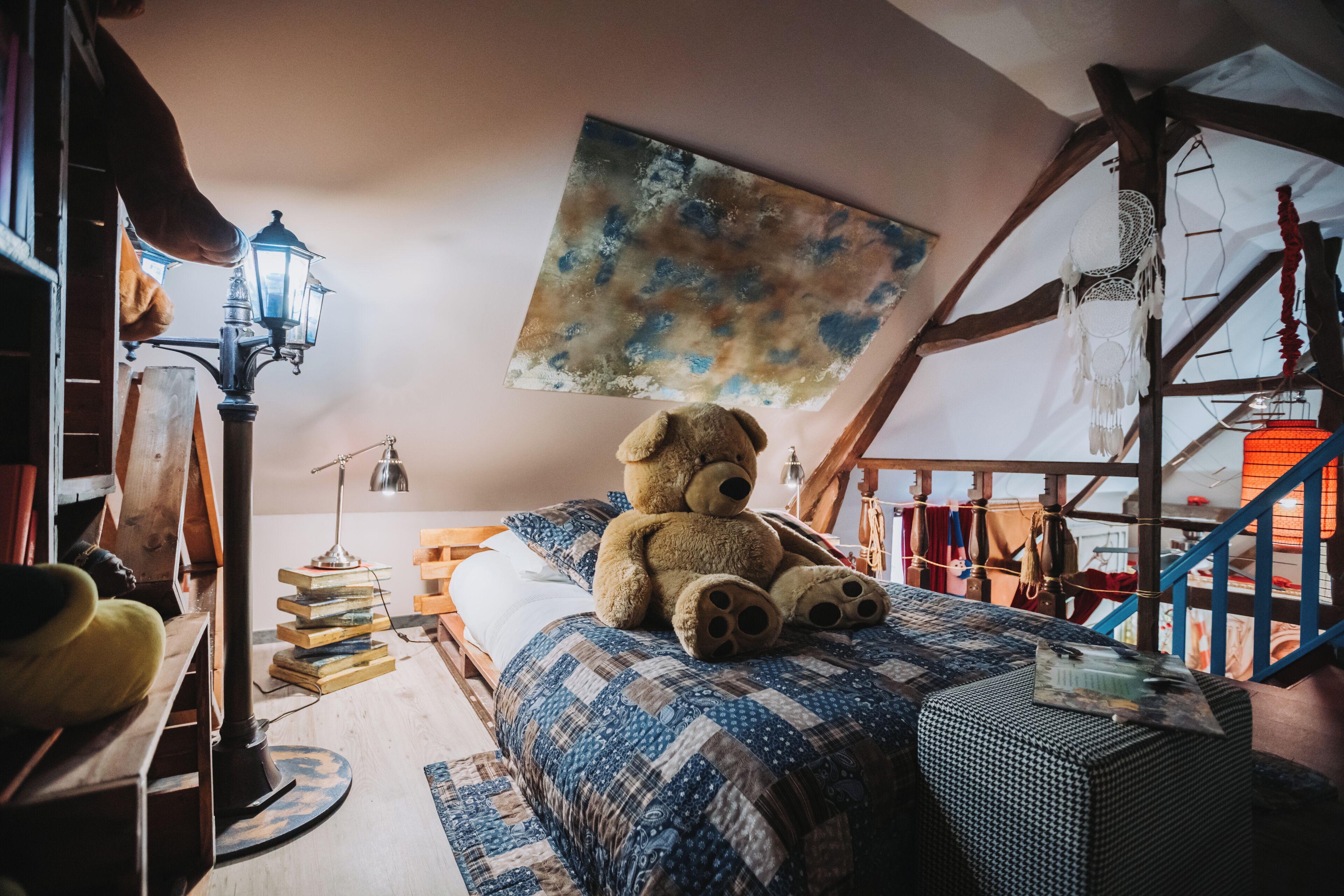 Acogedor dormitorio en ático con un gran oso de peluche sobre la cama, una escalera de madera y un cuadro abstracto colgado.