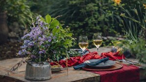 Mesa al aire libre para dos personas, con una maceta de metal con flores, dos copas de vino y un mantel rojo.