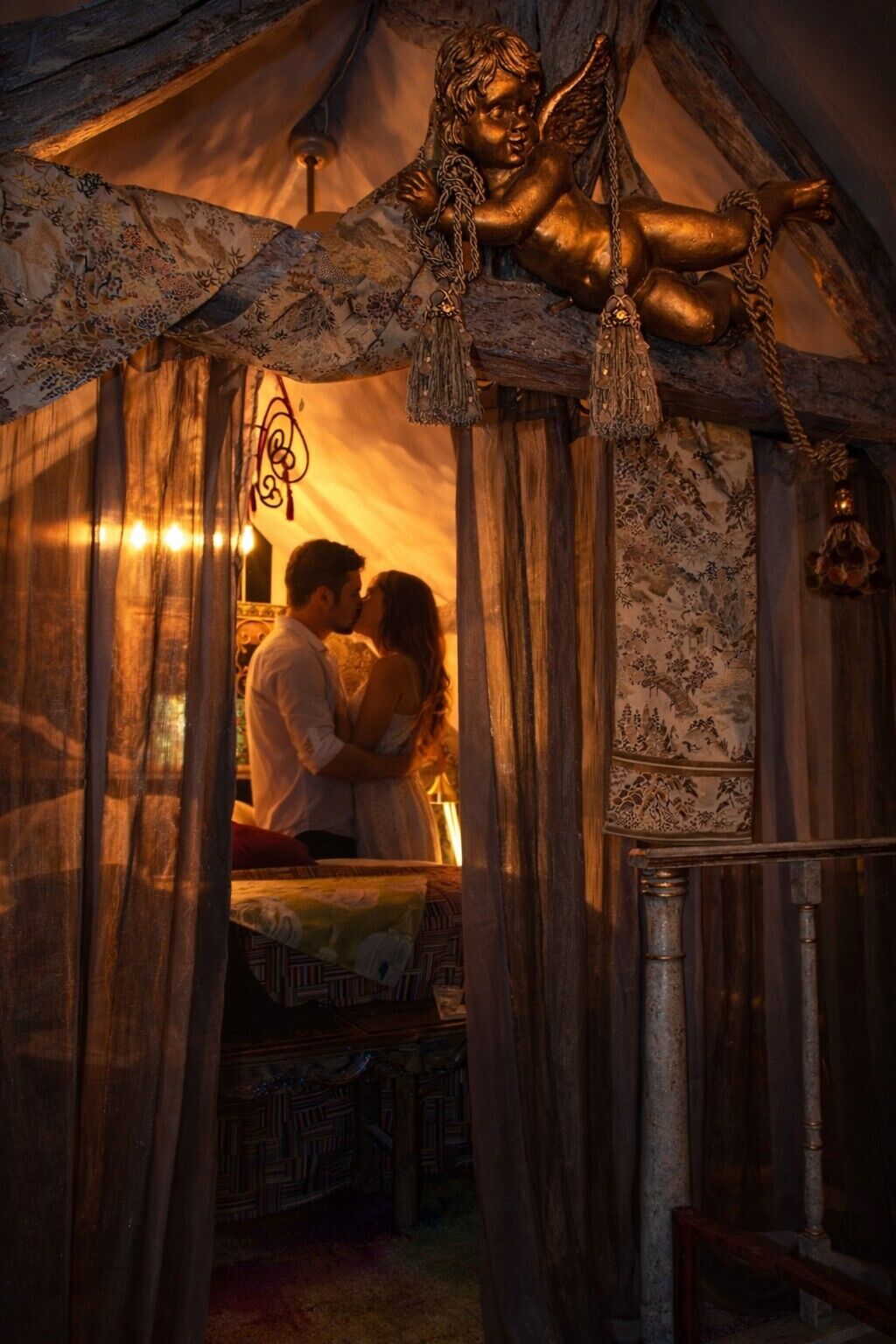 A tent-like structure with a decorative angel figure above, a patterned curtain, and a couple kissing.