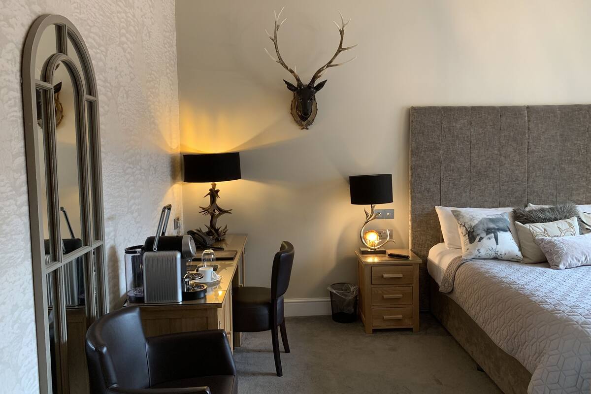 A bedroom with a bed, two bedside tables with lamps, a desk with a chair, a mirror, and a wall-mounted deer head.